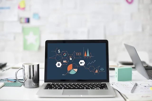 A laptop (MacBook) open on a cluttered desk, displaying a screen with many charts on it. The background has a wall with many papers pinned to it. SEO tools reporting dashboard: how to structure metrics and evidence